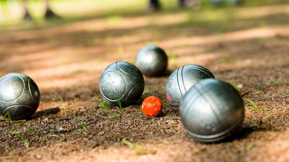 Gleise mit silbernen Boulekugeln und einer roten Kugel auf einem erdigen Boden in einem Park.