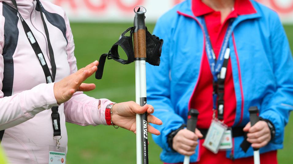 Zwei Frauen stehen auf einer Wiese, eine zeigt auf einen speziellen Stock für Blinde mit Halterung.