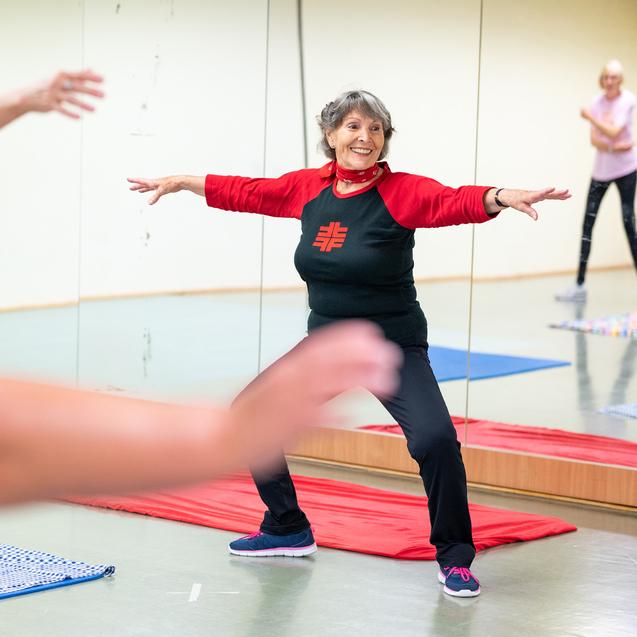Eine ältere Frau in Sportkleidung führt eine Dehnübung in einem Fitnessraum mit Spiegeln aus.