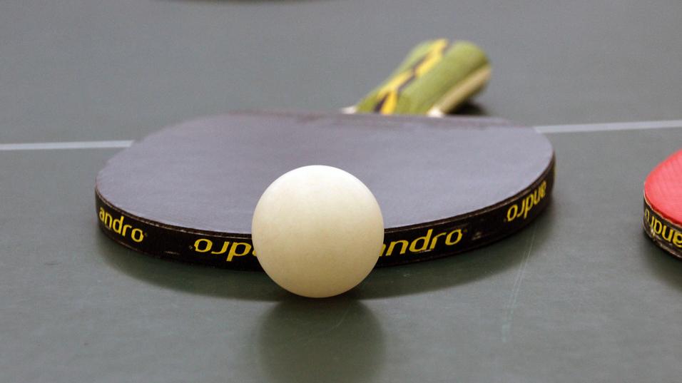Nahaufnahme eines Tischtennisschlägers mit weißem Ball auf einer grünen Tischtennisplatte.